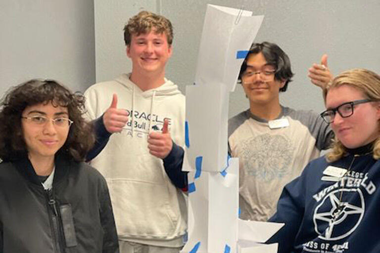 Four students stand around a tall paper structure held together with blue tape