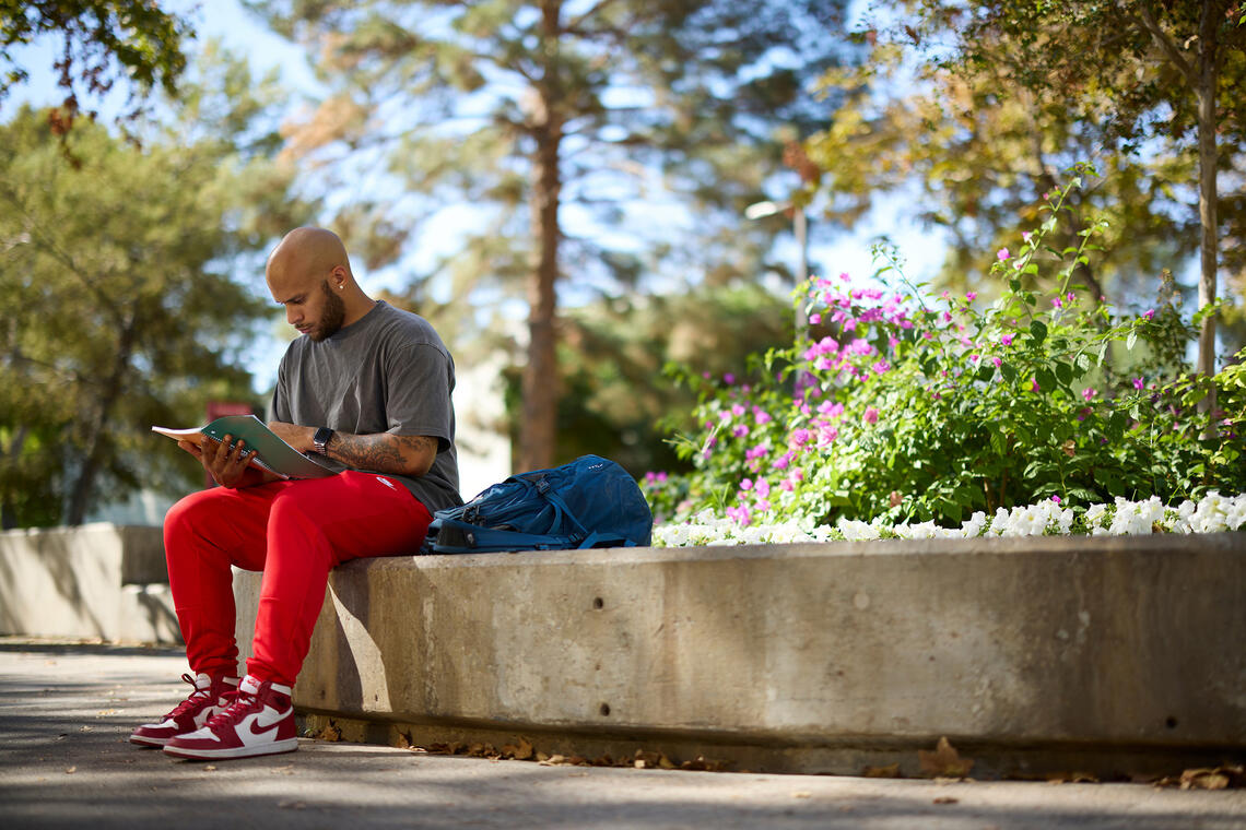 A student sitting on a low concrete wall outdoors, looking at a notebook.