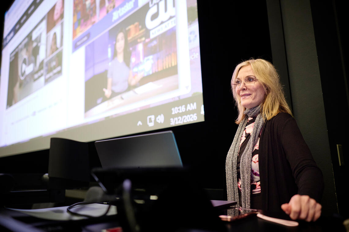 Photo of blonder woman in glasses at a podium teaching a class