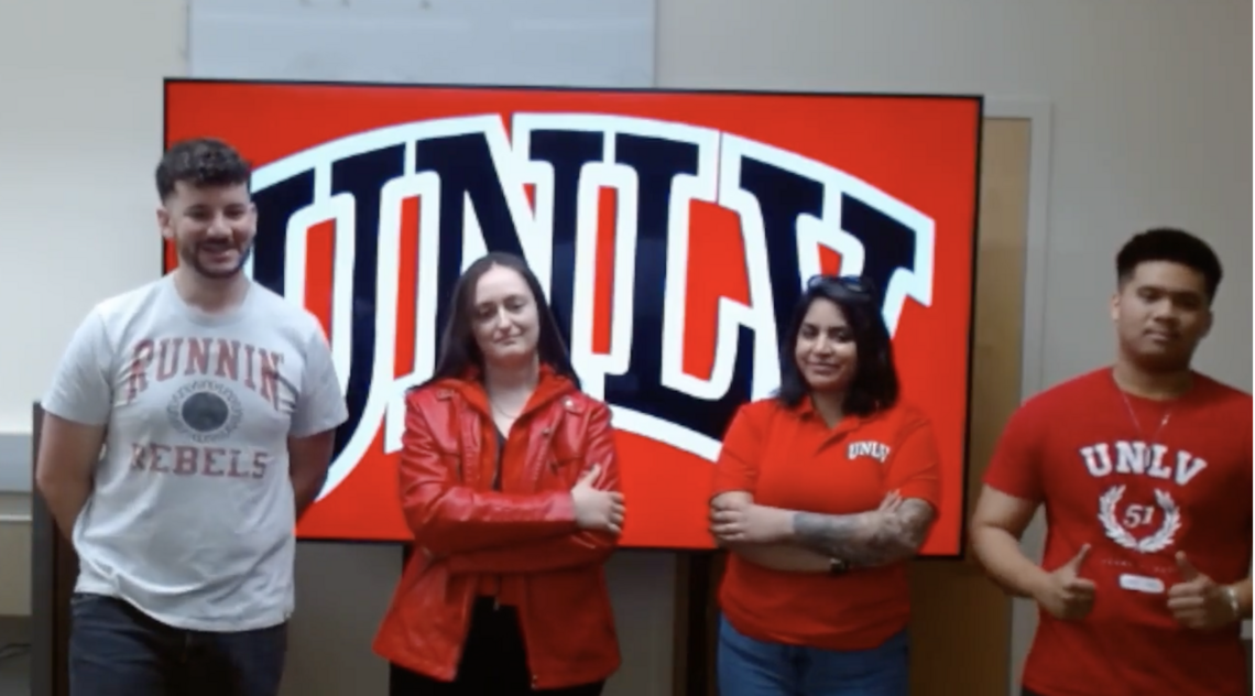 Members of Team Rebel Recon from UNLV stand in front of a large screen displaying the UNLV logo. From left to right are Luke Rasmusson, Bianca Ionescu, Shanzeh Aslam, and Hareign Casaclang.