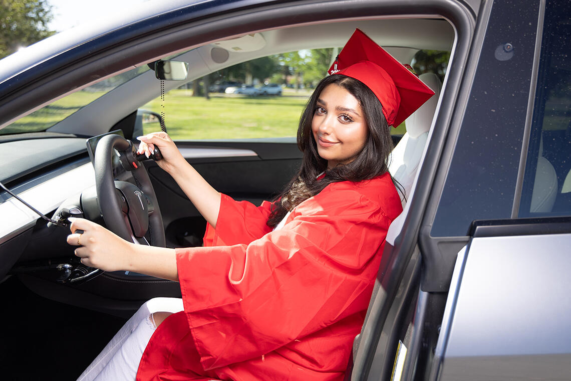 U.N.L.V. graduate wearing her red cap and gown sitting in the driver's seat of a vehicle