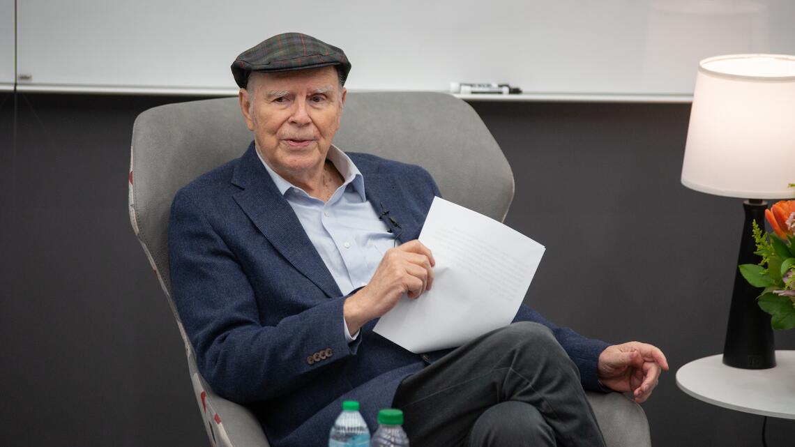 Norman McCombs, 2013 National Medal of Technology and Innovation recipient, sits holding papers during a fireside chat, wearing a plaid flat cap and navy blazer