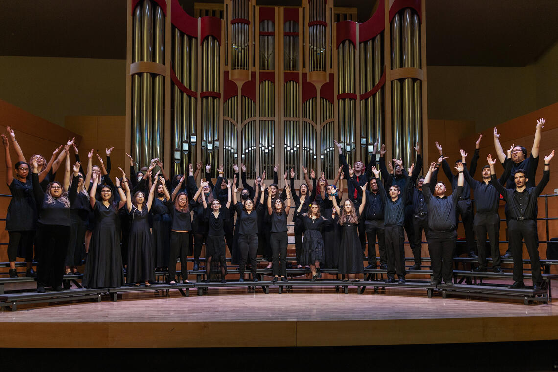 A dark stage with a crowd of choir singers with their hands raised on stage