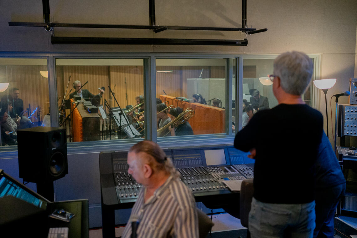 Aperson standing inside of a recording room as a band plays in the other room