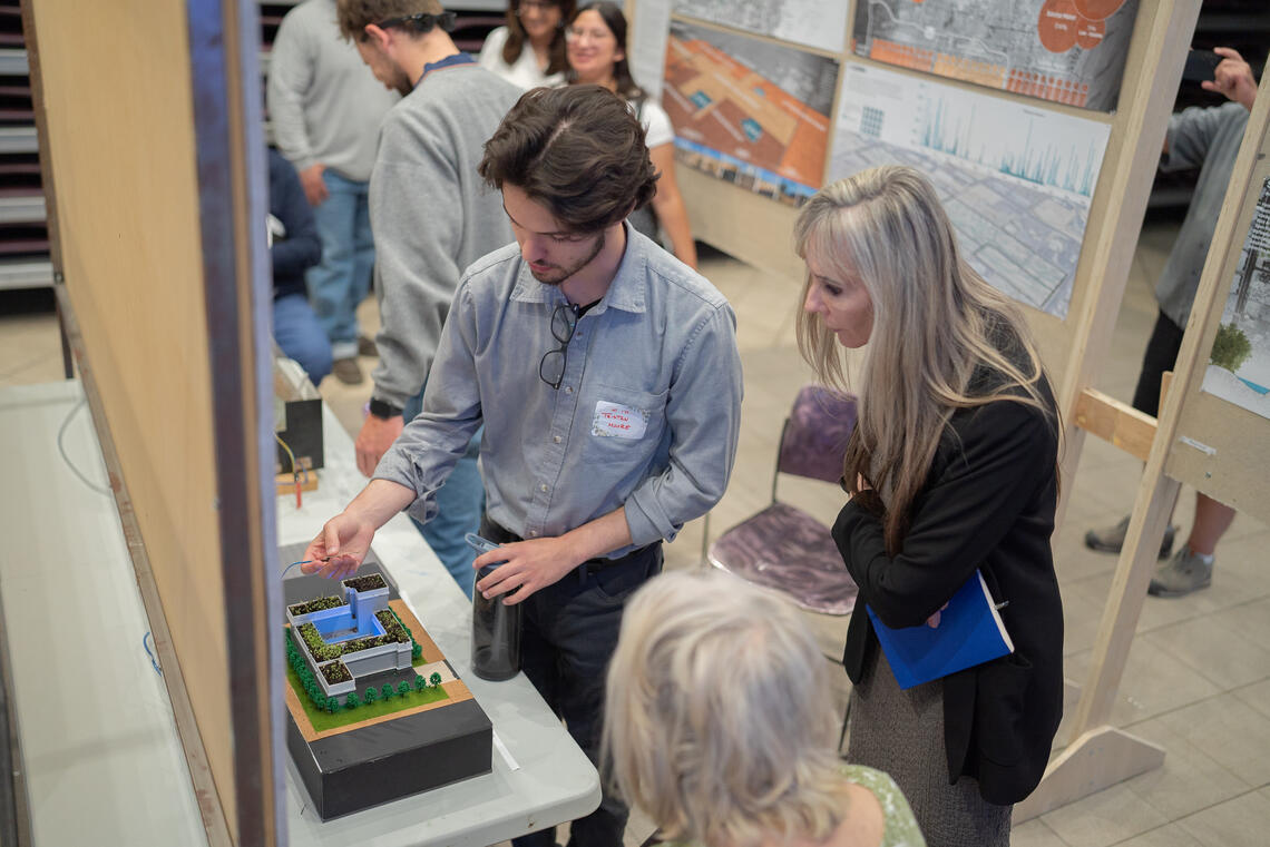A student with a name tag reading "Hi I'm Tristan Moore" showing 2 people a model of a two story modern house with greenery on the roof