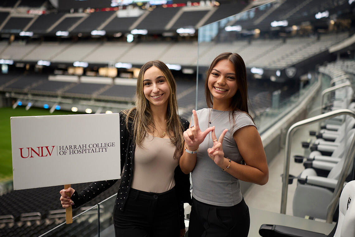 Two students smiles while standing inside of Allegiant stadium, one holding a sign reading "UNLV Harrah College of Hospitality"