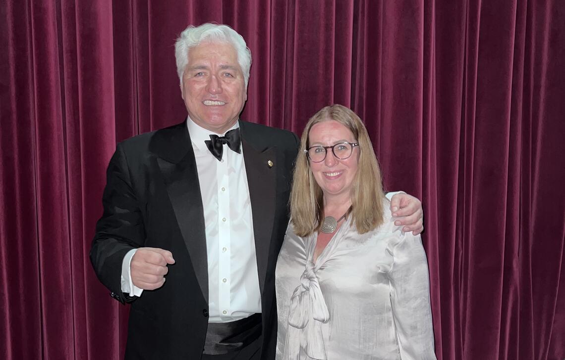 A man with white hair and a woman with glasses stand in front of a theatre curtain smiling big