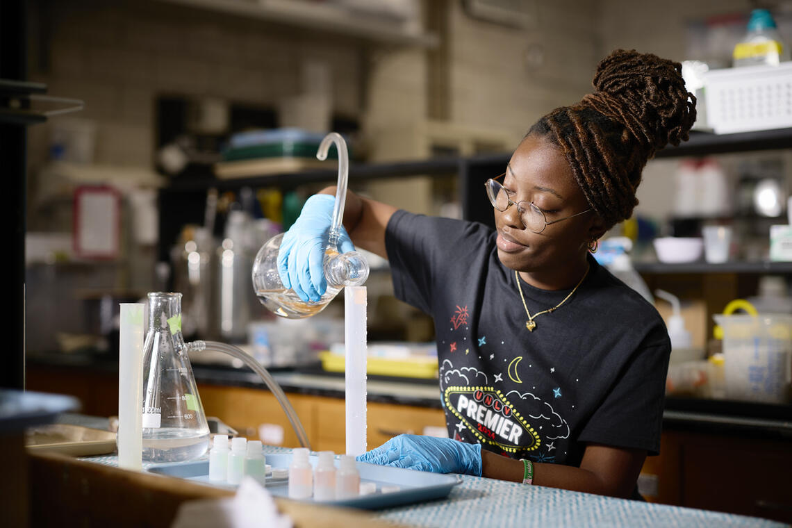 Woman in a black t-shirt and blue gloves pouring liquid into a beaker
