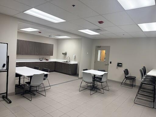 A small room with tan tiles and cream walls with 3 white tables with chairs on the ground and a station with grey wood cabinets and counters