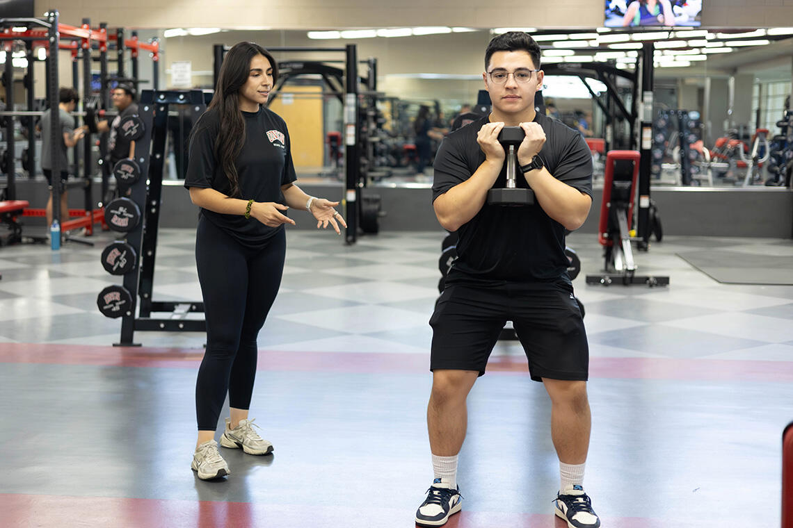 An fitness instructor besides a trainee who's lifting a dumbbell with two hands