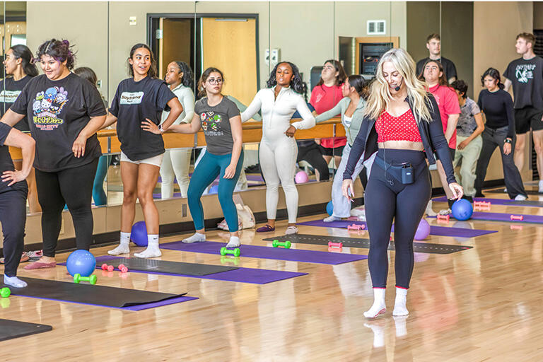 A fitness instructor leads a group of participants in an exercise class in a mirrored studio with yoga mats, dumbbells, and exercise balls.