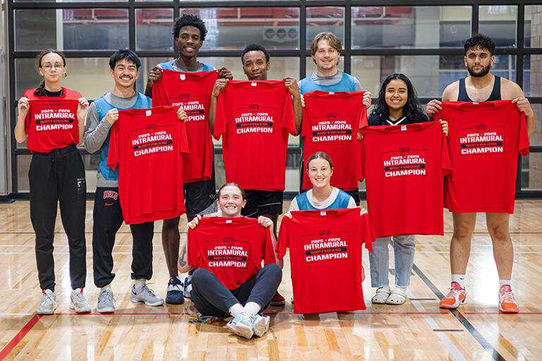 A group of nine athletes posing for photo on the court while holding up red championship t-shirts.