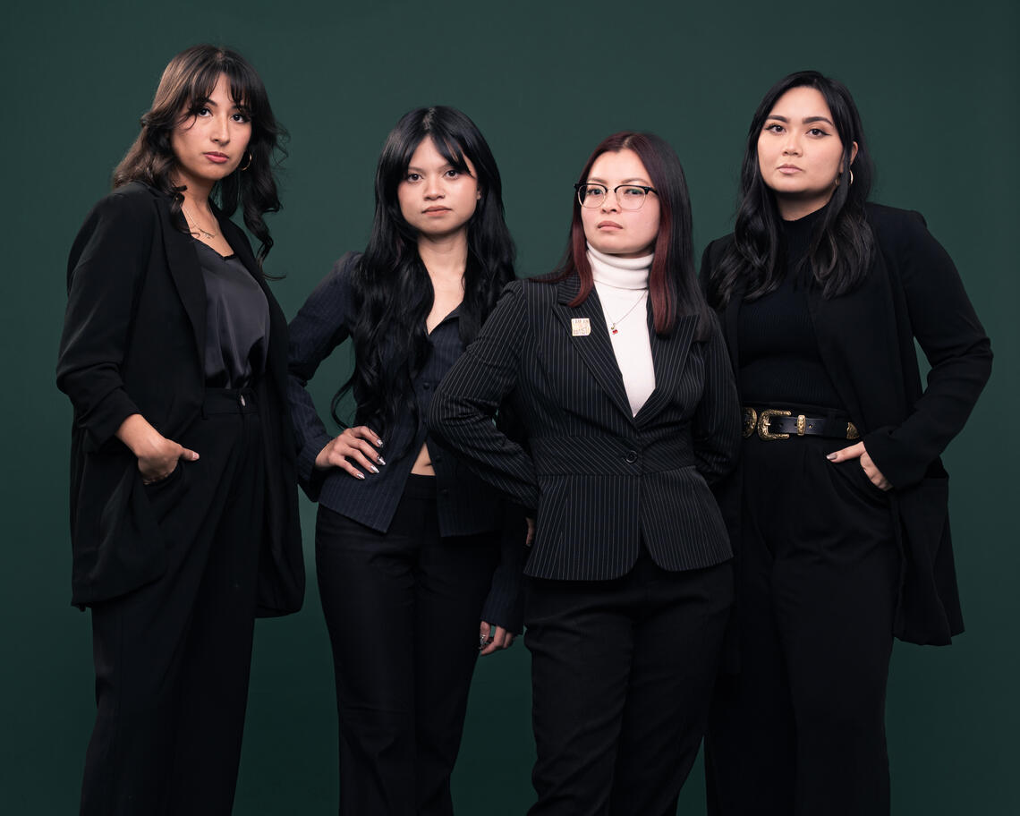 Four women in suits posing for the camera against a dark green background