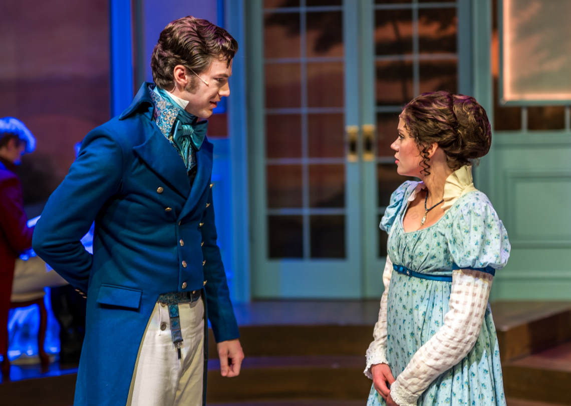 Two actors on stage for a play. Man in a navy blue suit, woman in a baby blue dress. They are speaking to each other.