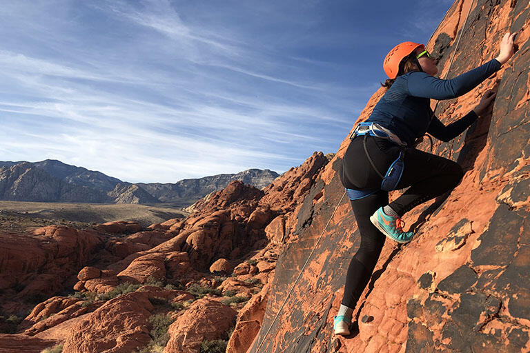 A rock cimber in climbing harness and helmet being belayed by rope as they scale up a rock face.