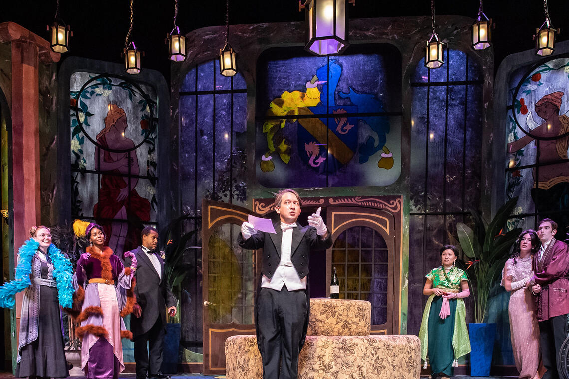 Scene during a play in which a group of well-dressed people surround a man in a tuxedo and listen with curiosity