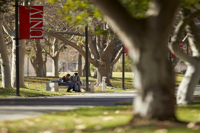 Wide shot of two students sitting on a bench that is located underneath the UNLV flag