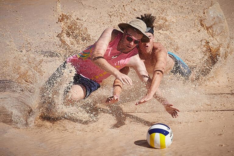 two male students dive through the mud towards a volleyball