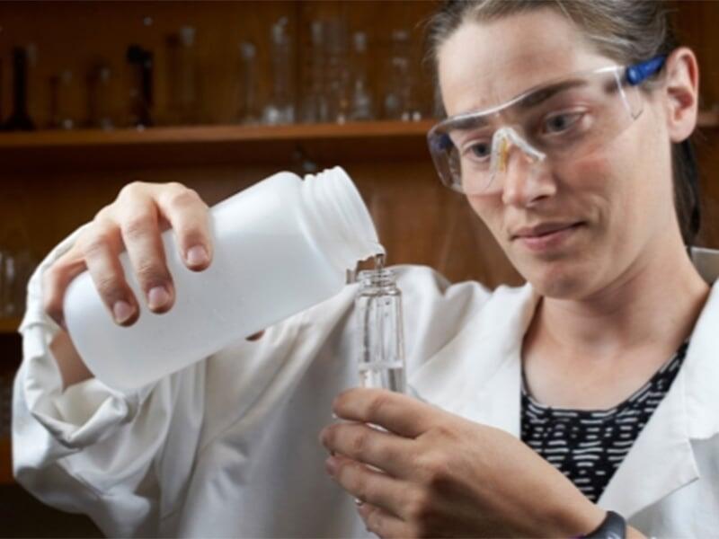 Female wearing safety goggles and a lab coat is pouring liquid form a white container into a clear tube shaped container
