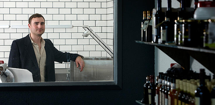 A man having his arm perched on a desk with the foreground showing a shelf of various alcohol bottles