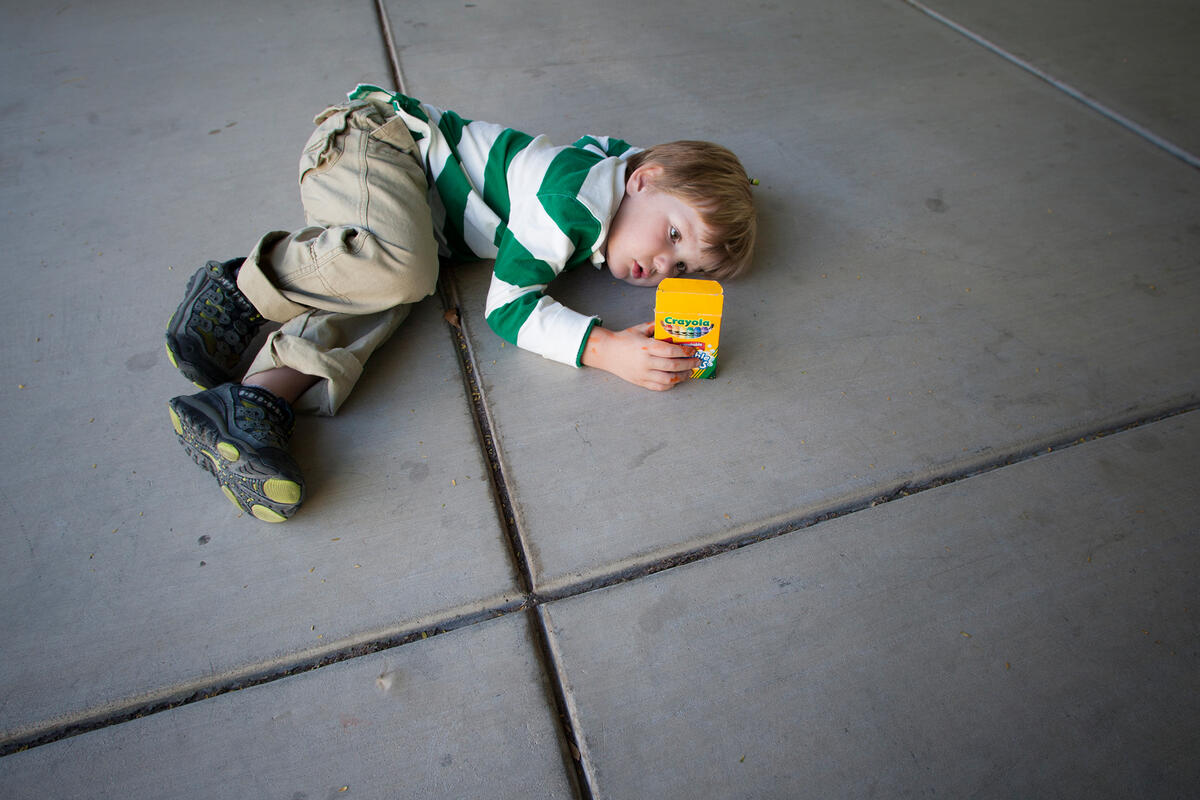 boy lays on the ground while looking at a pack of crayons