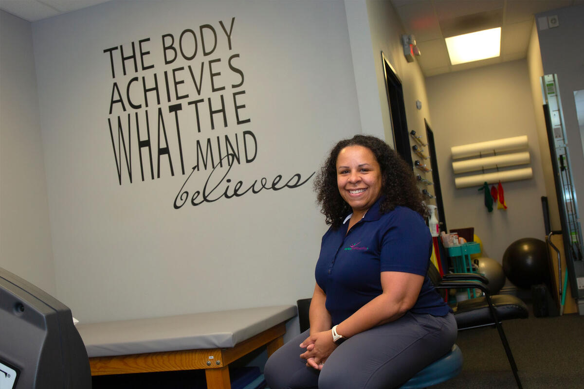 A woman sits in front of a sign reading "The Body Achieves What the Mind Believes"