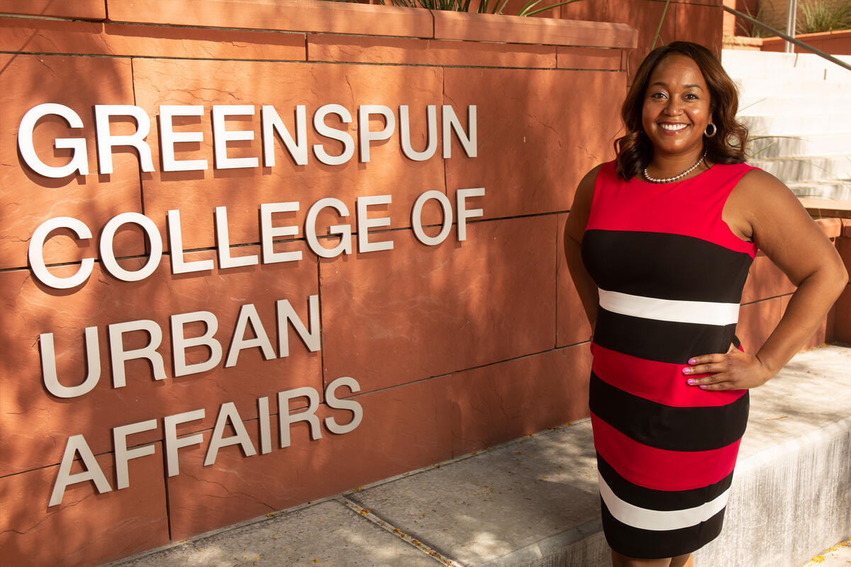 A woman in a red and black dress stands next to the College of Urban Affairs sign