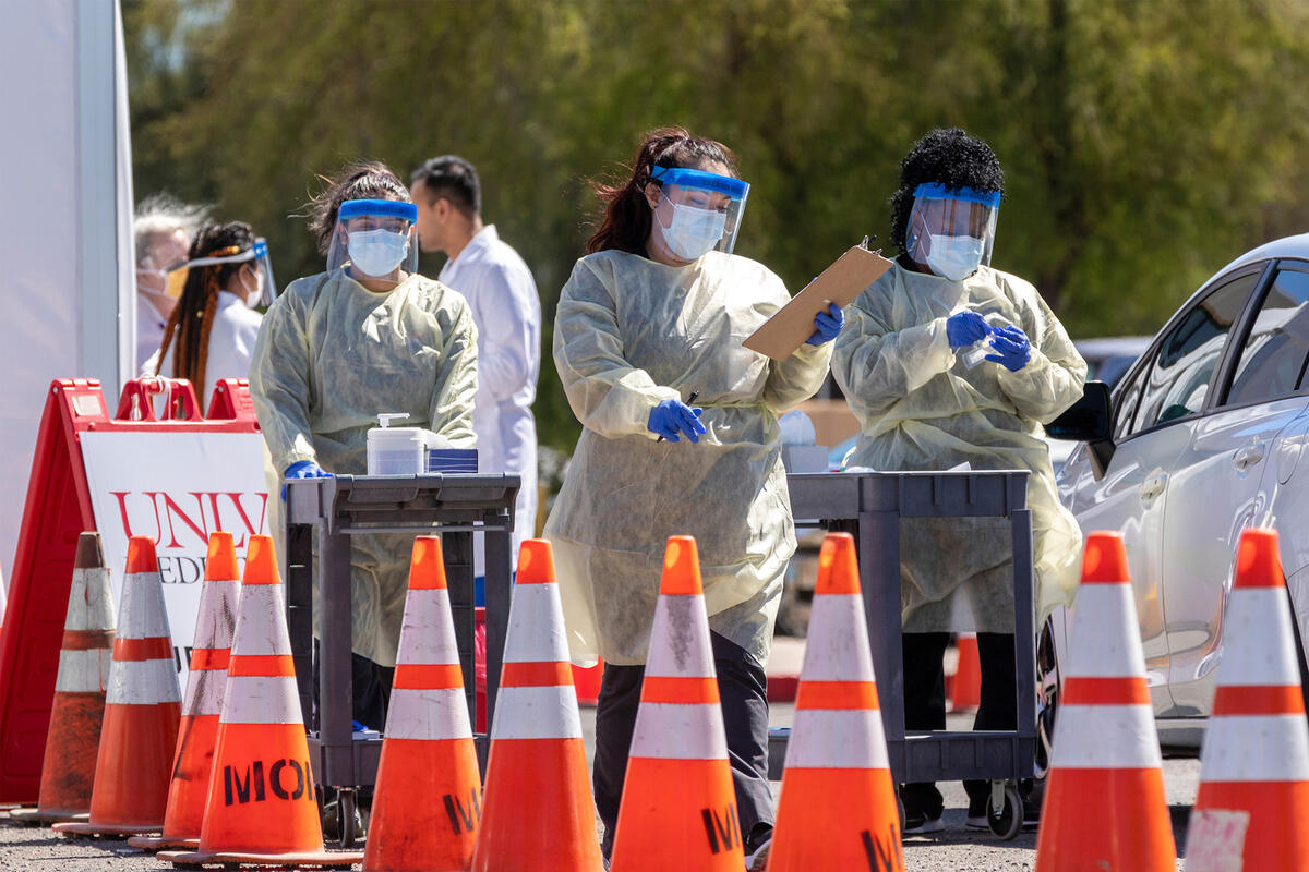 A group of health officials in protective equipment wait for al ine of cars passing orange cones