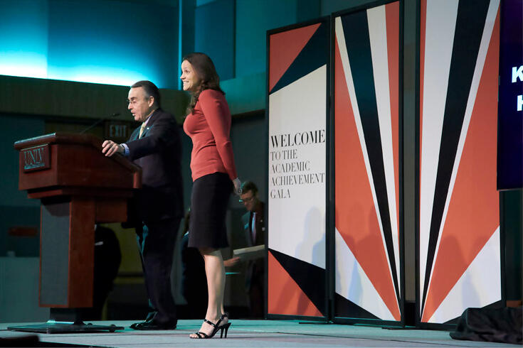Two people standing on stage while one speaks into the mic at a podium.