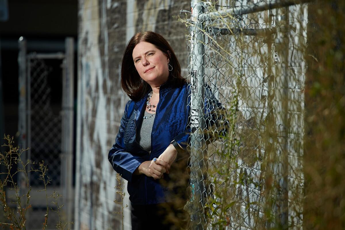 Alexis Kennedy leans against a chain-linked fence with brick wall in the background
