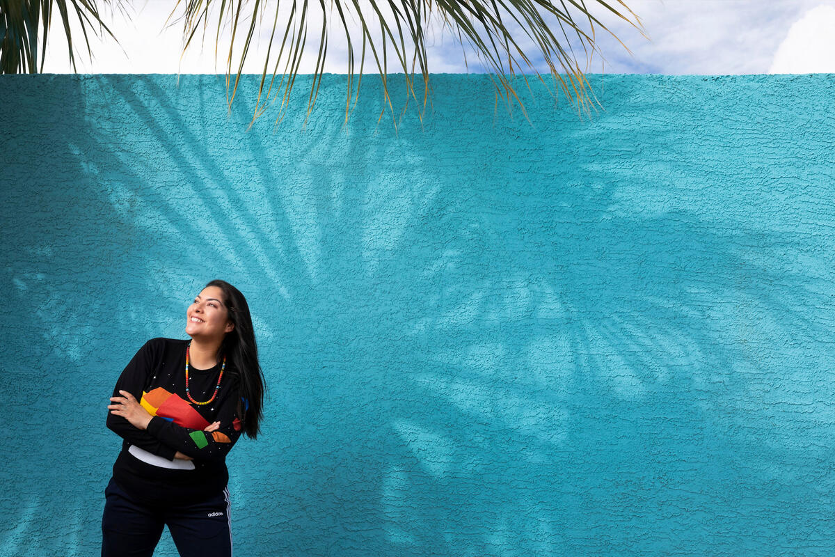 A woman stands next to a turquoise blue wall in the shadow of palm trees
