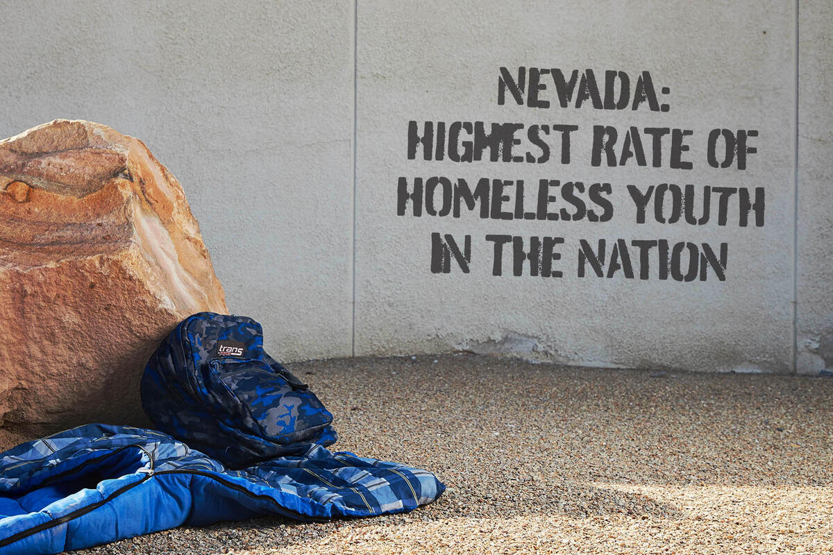 backpack and sleeping bag next to wall with "Nevada: Highest Rate of Homeless Youth in the Nation" spray painted on it