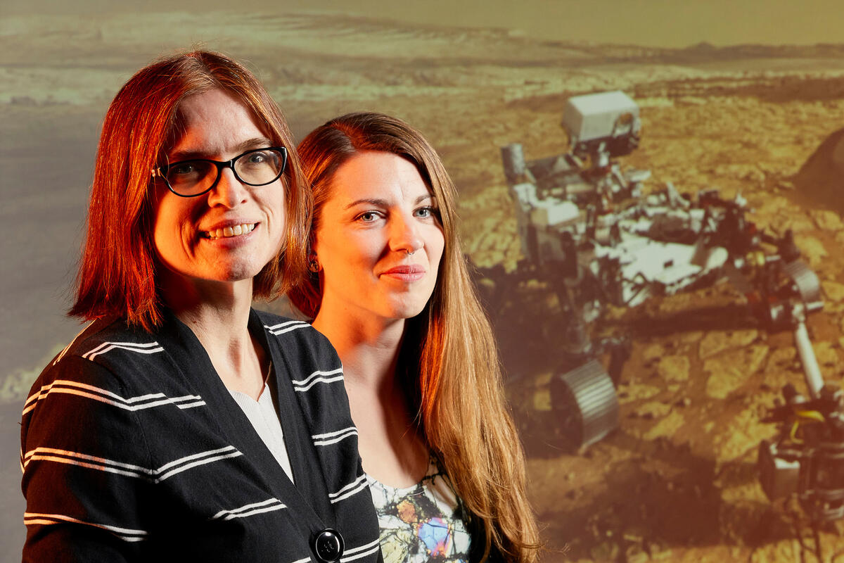 Two women stand in front of an image of a Martian rover