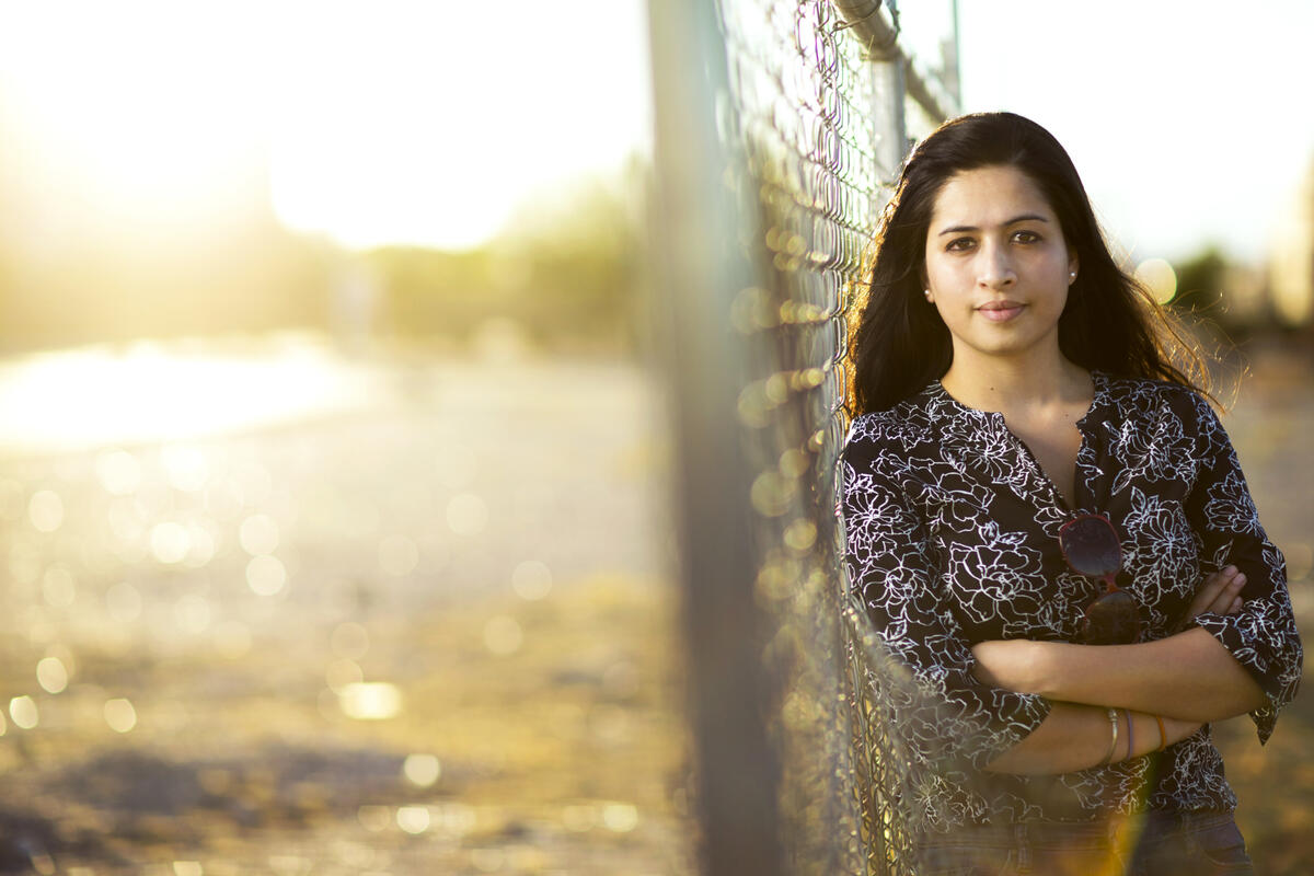 A woman leans against a chain-link fence as the sun sets in the background.