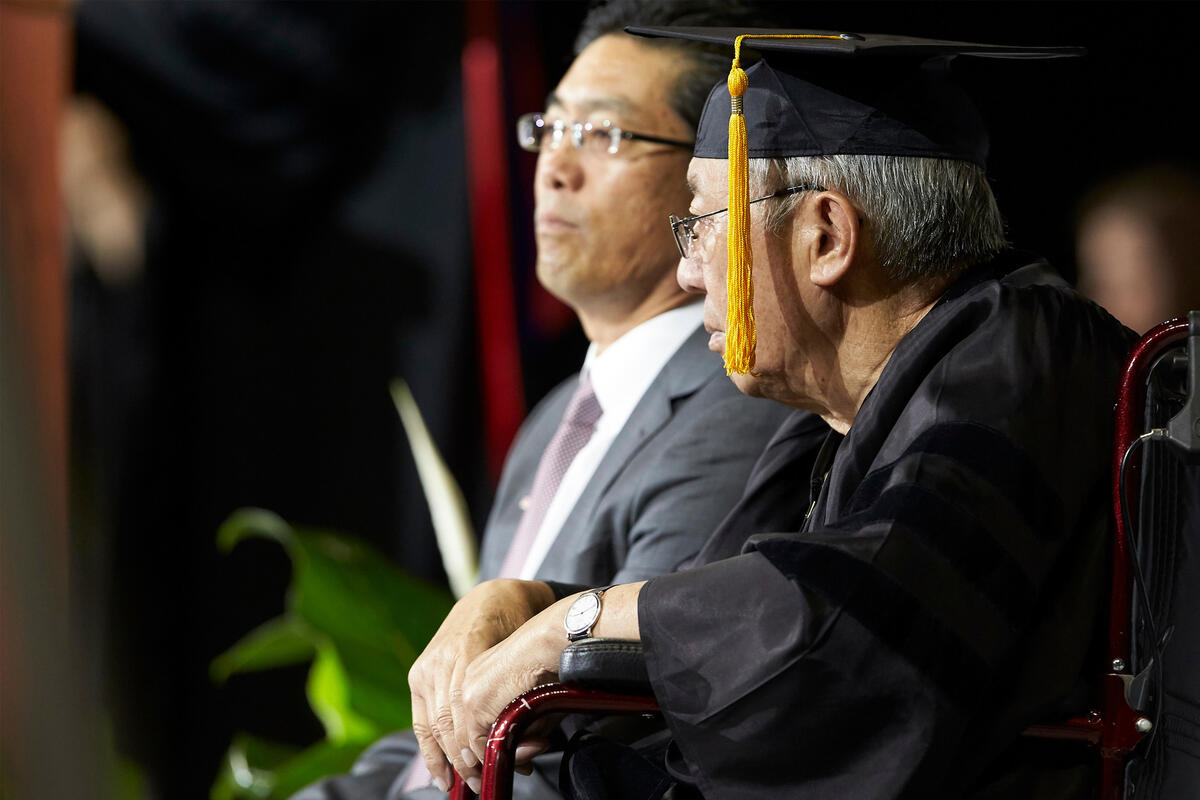 A man in a gown and mortarboard sits flanked by his son