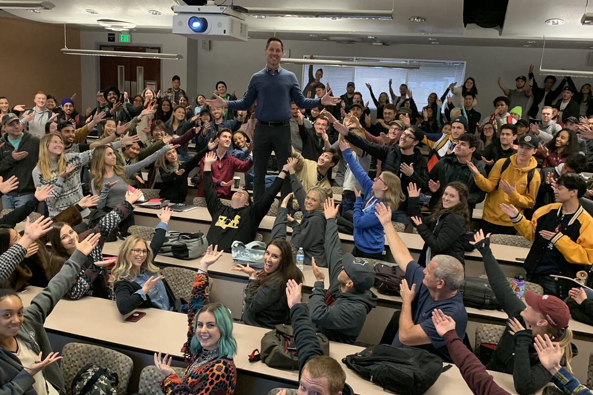 A photo of Danny Siciliano standing on a table with students pointing at him.