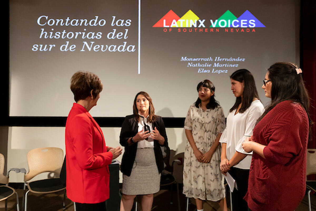 Group of five women standing in front of a screen. The screen has a slide from a presentation about Latin-x voice in southern Nevada.