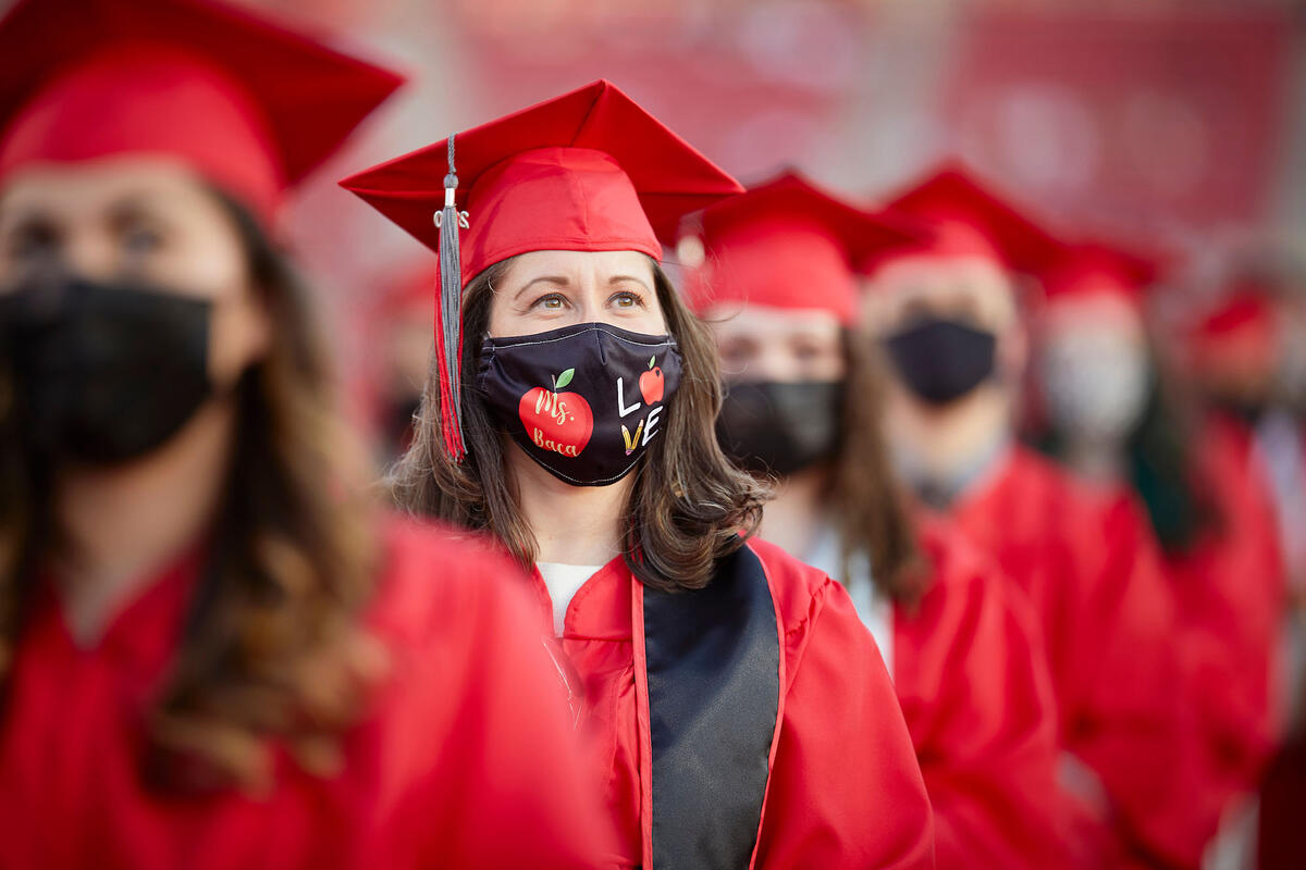Young woman wearing a red cap and gown and a teacher-themed mask that says Ms. Baca and Love