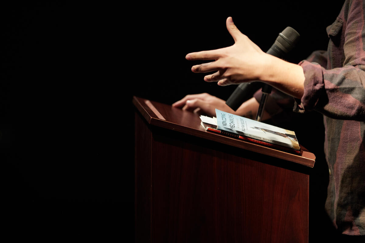 a person stands at a lectern with a microphone reading passages from a book