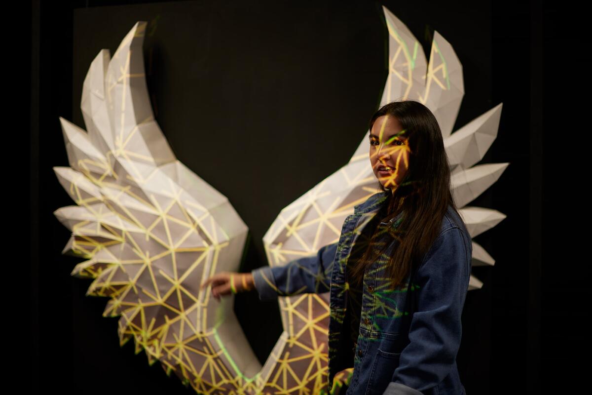 Student stands of fron a pair of wings with light projected on it.