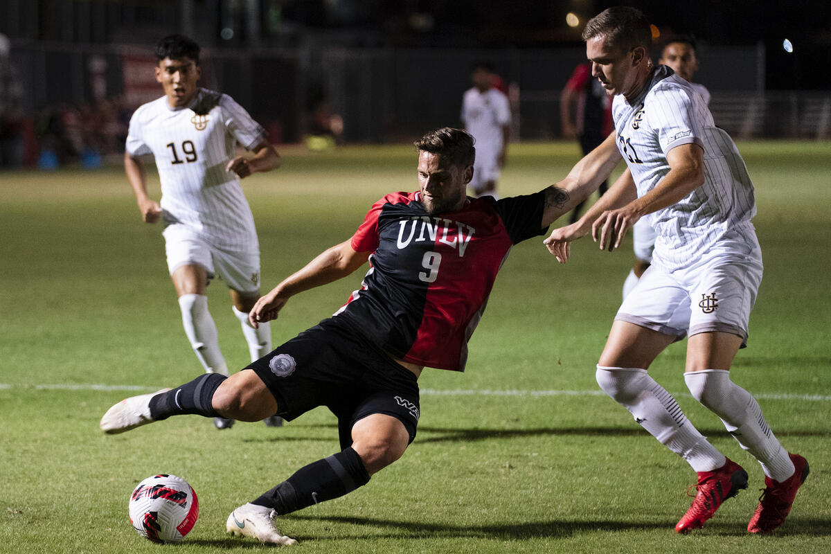 Three male players on soccer field mid game, UNLV player kicking the ball