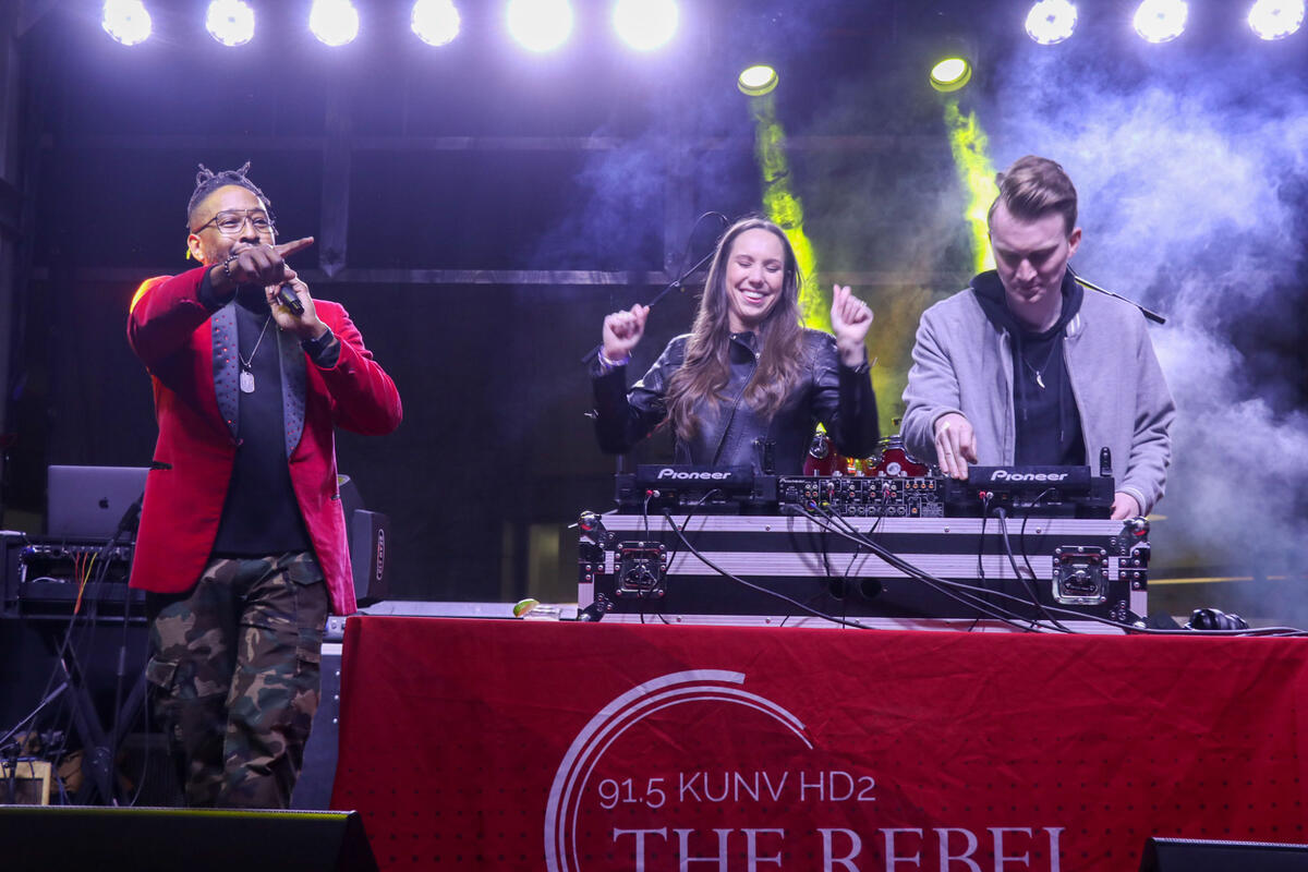 On stage, person on the left holding up a microphone and two people at a dj booth on the right