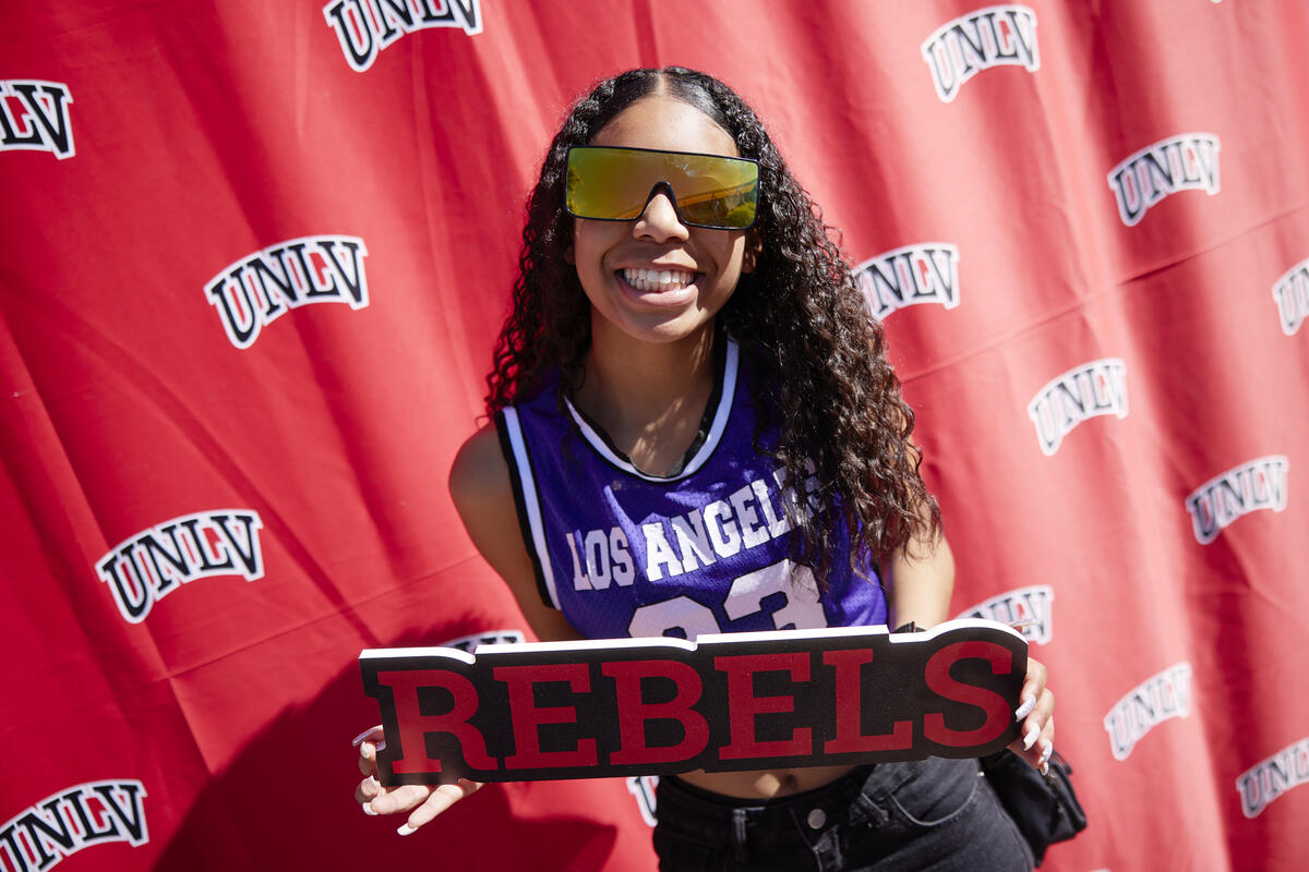 A woman wearing sunglasses who holds a sign that says &quot;REBELS&quot; standing in front of a backdrop with the word &quot;UNLV&quot; repeated multiple times.