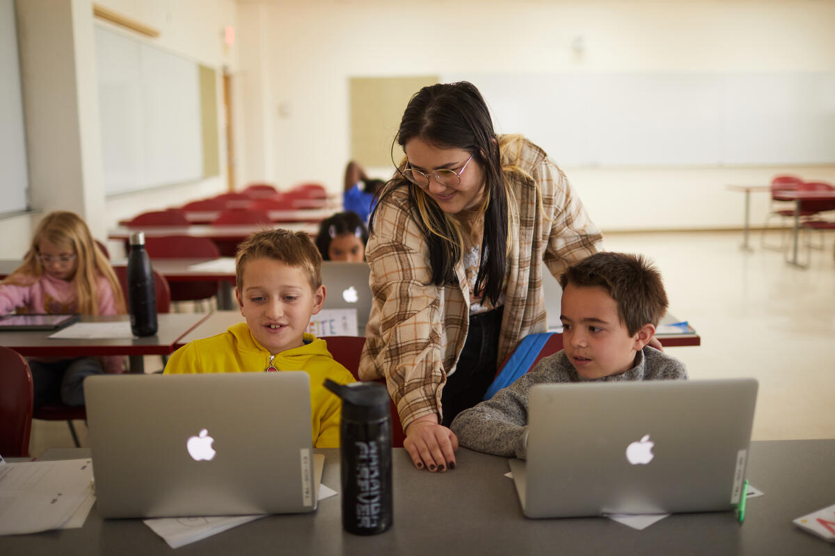 Instructor assisting two young children who are working on laptops.