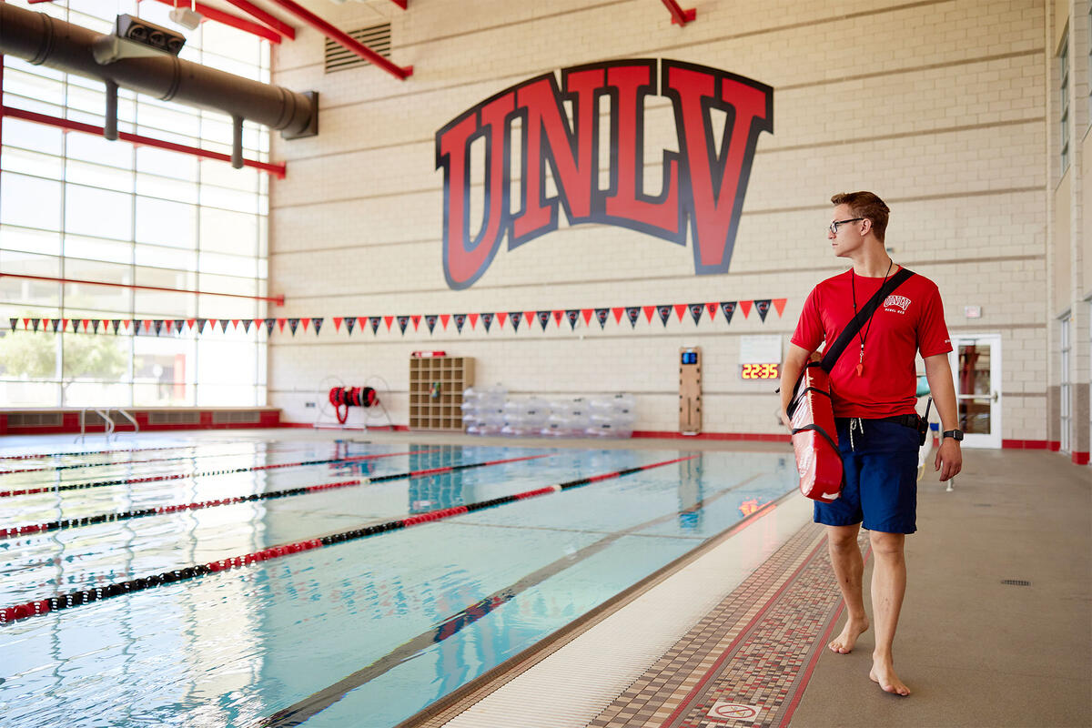 A UNLV lifeguard waking along the outer edge of a large indoor pool