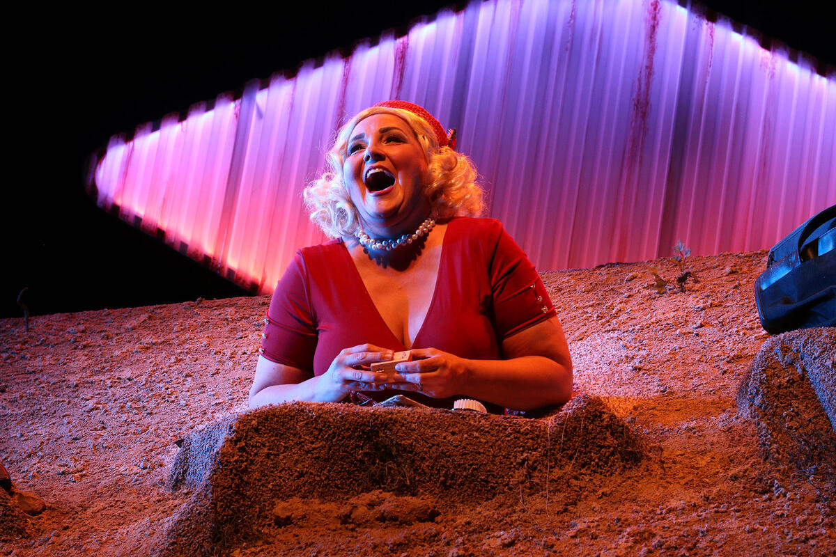 Woman with a red hat, blonde wig, and red shirt singing with a huge smile on her face.