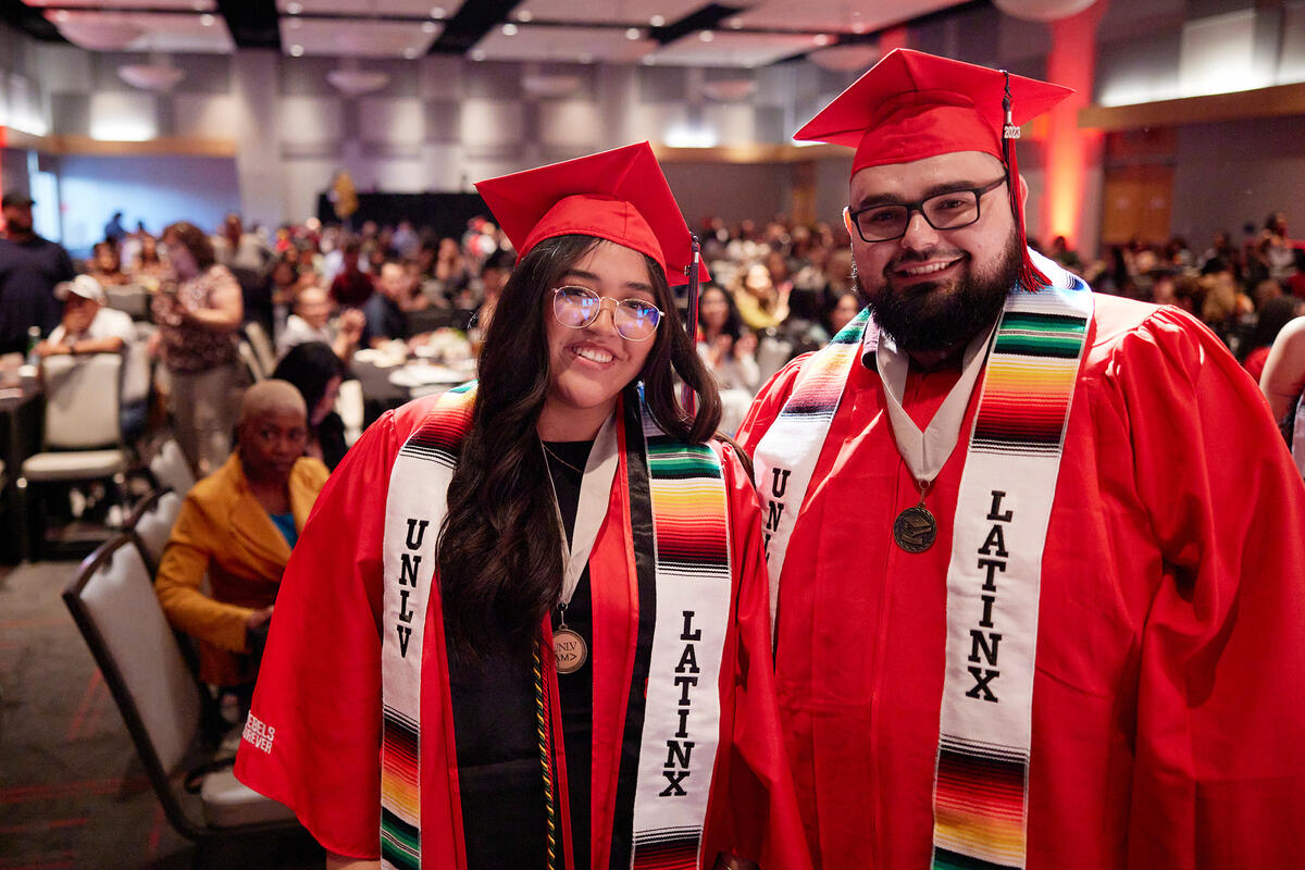 Two Latinx graduates wearing red commencement caps and gowns and the UNLV Latinx sashes