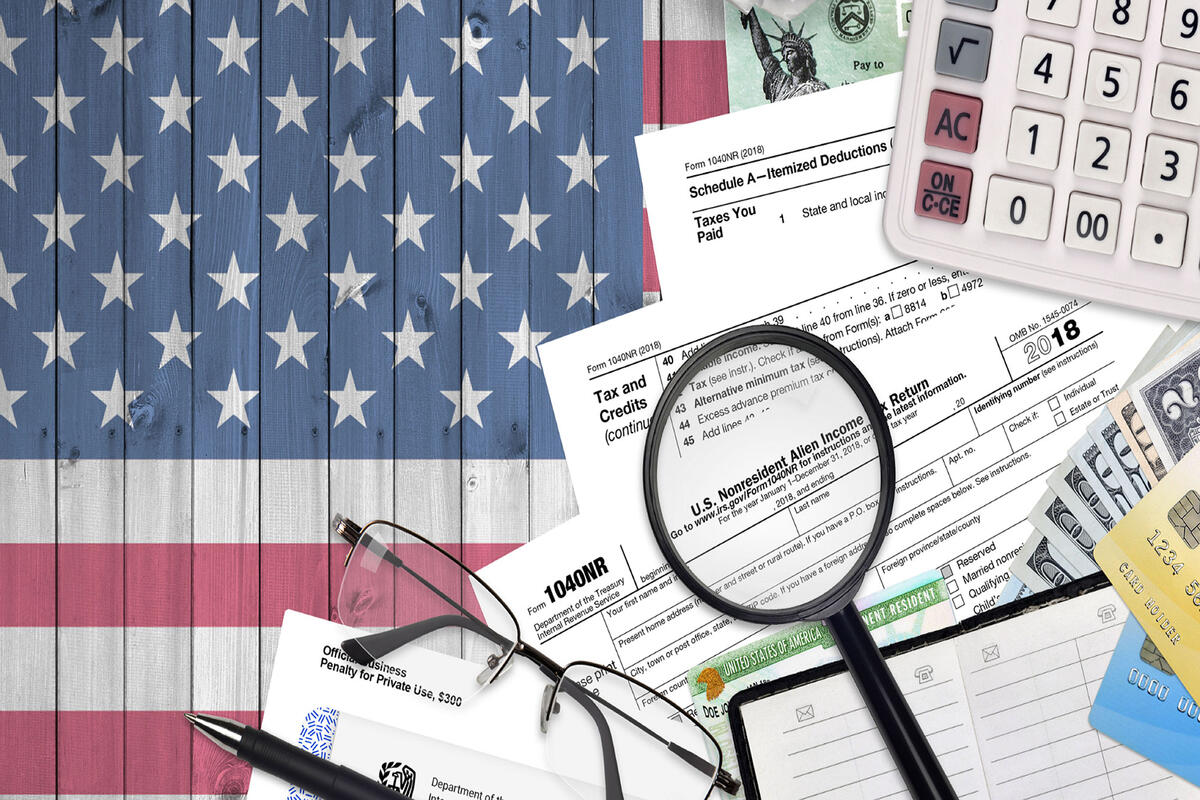 A graphic of the USA flag in the background and a pile of tax related documents, a magnifying glass, USA dollars, and a calculator