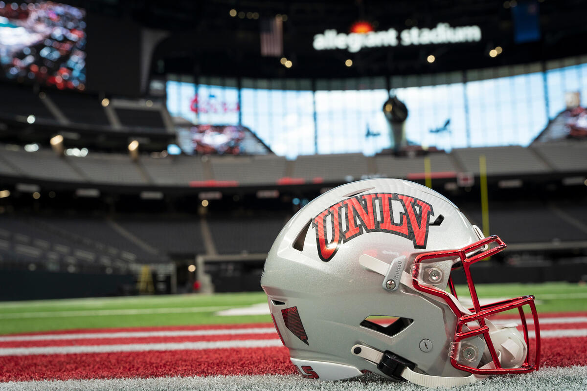 a UNLV football helmet placed on the turf with an Allegiant Stadium sign and empty bleachers visible in the background