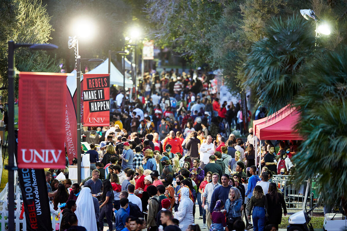 large crowd moving through the academic mall on the UNLV Campus during homecoming. Various booths and banners are set up on either side of the walkway.